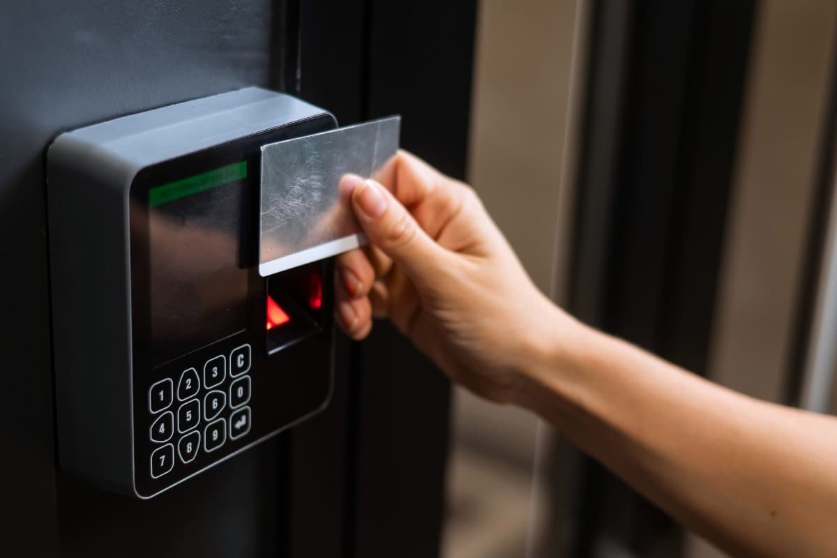 A hand presenting an access card to a wall-mounted reader with keypad and fingerprint sensor
